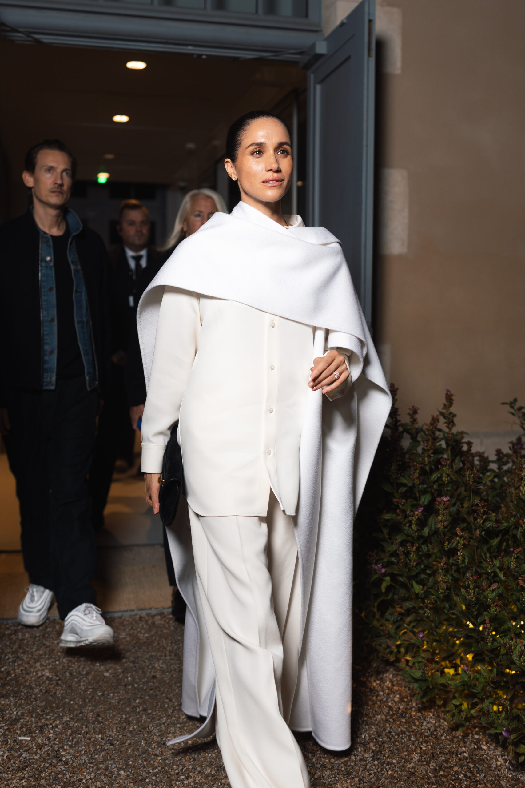 Meghan Markle in an all-white outfit with a flowing shirt, wide-leg trousers, and a dramatic cape, arriving at Paris Fashion Week.